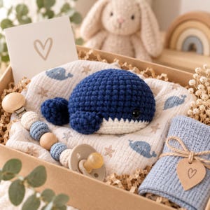 May include: A gift box containing a dark blue crocheted whale, a white muslin swaddle with blue whale and star prints, a pacifier clip with wooden and crocheted beads, a light blue textured cloth with a heart tag, and a card with a heart drawing.