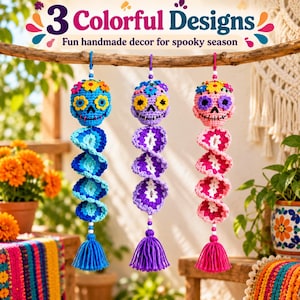 Può includere: Tre ornamenti a forma di teschio all'uncinetto appesi a un ramo. Ogni teschio presenta accenti floreali colorati e una coda a spirale che termina con una nappa. I teschi blu, viola e rosa sono decorati con fiori realizzati in filato.
