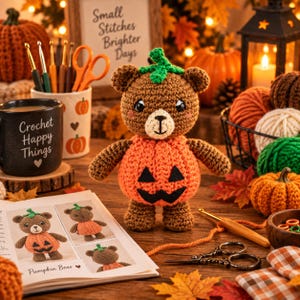 May include: A brown crocheted bear plush toy wearing an orange pumpkin-shaped body with a black felt jack-o'-lantern face and a green stem on its head. The bear has a cream-colored muzzle, embroidered facial features, and pink blush accents.