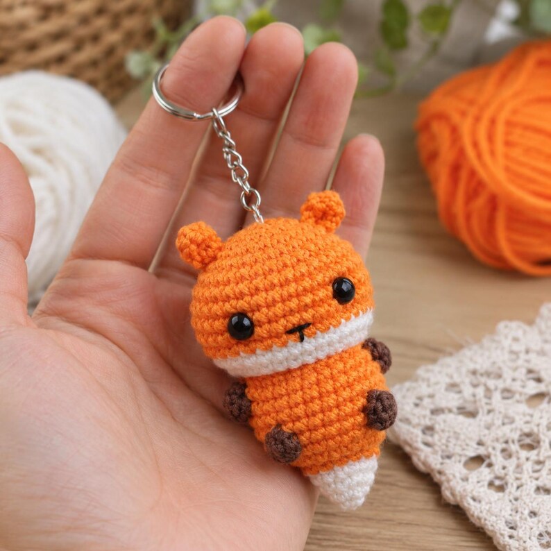May include: A small, handmade crochet fox keychain featuring bright orange yarn, a white muzzle, and a white-tipped tail. The fox has two small dark brown paws, black plastic bead eyes, and a simple embroidered nose, attached to a silver metal ring.