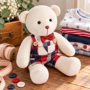 May include: A cream-colored plush teddy bear wearing red, navy blue, and white plaid overalls with a matching red bow tie. The bear has black button eyes and a stitched red nose. The overalls feature two small wooden buttons at the straps.