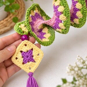 K&ouml;nnte beinhalten: Eine spiralf&ouml;rmige H&auml;kel-H&auml;ngedekoration aus gr&uuml;nem, gelbem und violettem Garn. Die Struktur enth&auml;lt ein quadratisches Granny-Square-Motiv am unteren Ende, das mit einer violetten Perle und einer violetten Garnquaste an der Basis verziert ist.