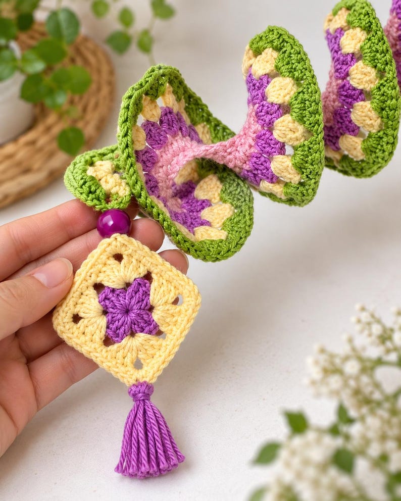 K&ouml;nnte beinhalten: Eine spiralf&ouml;rmige H&auml;kel-H&auml;ngedekoration aus gr&uuml;nem, gelbem und violettem Garn. Die Struktur enth&auml;lt ein quadratisches Granny-Square-Motiv am unteren Ende, das mit einer violetten Perle und einer violetten Garnquaste an der Basis verziert ist.