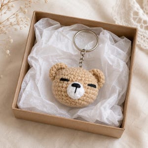 May include: A tan crochet bear head keyring featuring a white muzzle, black embroidered eyes, and a black nose. The bear head is attached to a silver-toned metal split ring by a short chain, resting inside a square brown cardboard gift box.