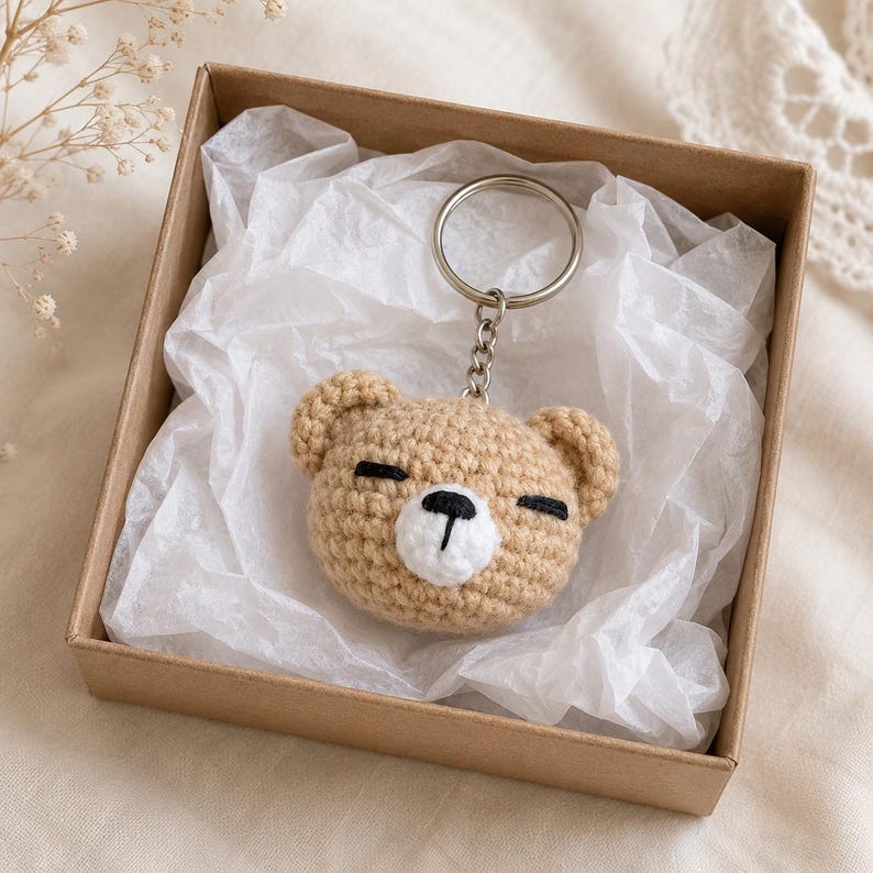 May include: A tan crochet bear head keyring featuring a white muzzle, black embroidered eyes, and a black nose. The bear head is attached to a silver-toned metal split ring by a short chain, resting inside a square brown cardboard gift box.