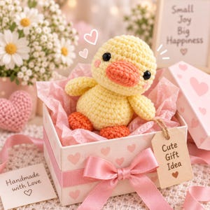 May include: A yellow crocheted duck plush with black eyes and an orange beak and feet, sitting inside a white gift box decorated with pink hearts. The box is tied with a pink ribbon and features a brown tag labeled Cute Gift Idea.
