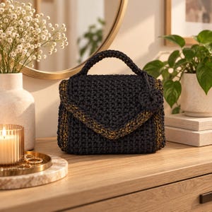 Puede incluir: Un bolso de ganchillo en color gris carb&oacute;n oscuro con un asa superior estructurada y cierre de solapa. El bolso presenta detalles en hilo dorado met&aacute;lico tejido a lo largo de los bordes de la solapa y los laterales, e incluye un peque&ntilde;o adorno.