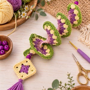 K&ouml;nnte beinhalten: Ein spiralf&ouml;rmiges H&auml;kel-H&auml;ngeornament mit gr&uuml;nen, gelben und violetten Garnsegmenten. Die Struktur besteht aus einer quadratischen Granny-Square-Basis mit einer violetten Quaste, akzentuiert durch violette Perlen und eine Schlaufe.