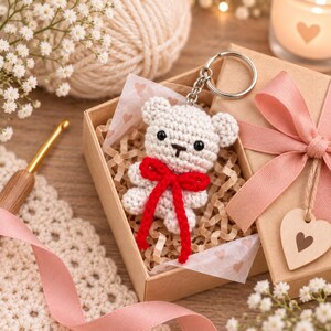 May include: A white crocheted bear keychain with black bead eyes and a small embroidered nose, featuring a bright red yarn bow tied around its neck. The bear is nestled inside a small cardboard gift box filled with shredded paper filler.