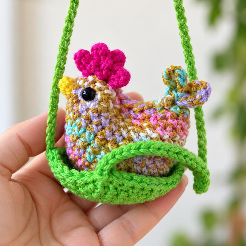 May include: A small, multicolored crochet chicken ornament featuring a bright pink comb, a yellow beak, and black bead eyes. The chicken sits inside a small, green crocheted basket that is suspended by two green crocheted straps.