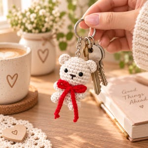 May include: A cream-colored crocheted bear keychain featuring black bead eyes, a small embroidered nose, and a bright red yarn bow tied around its neck. The bear is attached to a silver metal keyring that holds two standard silver metal keys.
