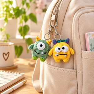 Peut inclure: Deux porte-clés monstres en crochet attachés à un sac à dos beige. Un monstre est vert avec des cornes jaunes et un œil, tandis que l'autre est jaune avec des cornes violettes et deux yeux. Les deux ont de petites dents blanches et des fermoirs.