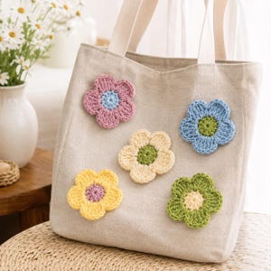 May include: A beige canvas tote bag featuring five crocheted flower appliques in various colors including pink, blue, yellow, and green. The bag has two sturdy white fabric handles attached to the top, providing a simple and functional design.