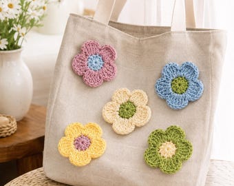 Op de afbeelding: Een beige canvas draagtas met vijf gehaakte bloemapplicaties in diverse kleuren, waaronder roze, blauw, geel en groen. De tas heeft twee stevige witte stoffen handvatten aan de bovenkant, wat zorgt voor een eenvoudig en functioneel ontwerp.