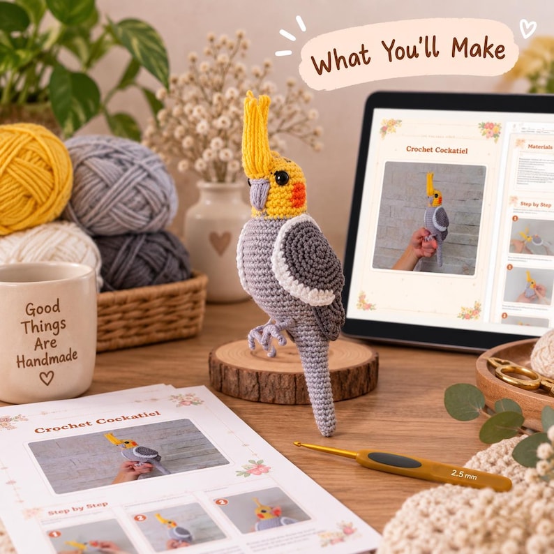 May include: A crocheted cockatiel bird with a yellow crest, grey body, and orange cheek patches, standing on a wooden slice. Beside it are a 2.5 mm crochet hook, yarn balls, a mug with the text Good Things Are Handmade, and a digital pattern.