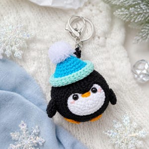 May include: A round, crocheted penguin keychain featuring a black body, a white belly, and orange feet. The penguin wears a bright blue crocheted beanie with a white pom-pom on top. It includes a silver-toned metal split ring and a lobster clasp.