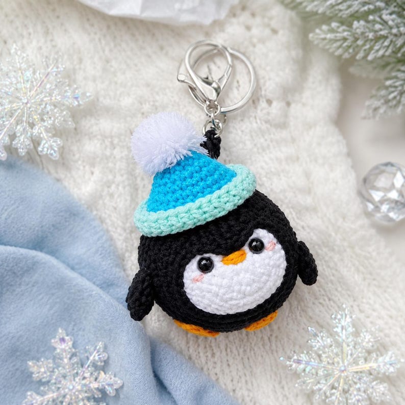 May include: A round, crocheted penguin keychain featuring a black body, a white belly, and orange feet. The penguin wears a bright blue crocheted beanie with a white pom-pom on top. It includes a silver-toned metal split ring and a lobster clasp.