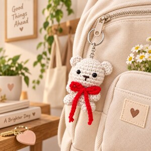 May include: A cream-colored crocheted bear keychain attached to a beige fabric backpack. The bear features black bead eyes, a small stitched nose, and a bright red yarn bow tied around its neck. The backpack has a small square patch with a brown heart.