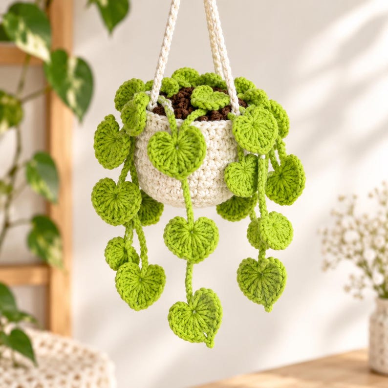 May include: A crocheted hanging plant featuring a cream-colored pot and vibrant green heart-shaped leaves on trailing vines. The plant is suspended by a thick, cream-colored crocheted cord, and the soil is represented by dark brown yarn.