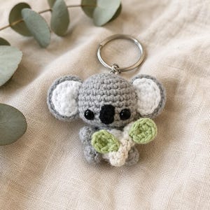 May include: A small, handmade crochet koala keychain featuring a grey body, white inner ears, and black plastic eyes. The koala has a black nose and is holding a small, light green and cream-colored crocheted eucalyptus leaf branch attached to a metal ring.
