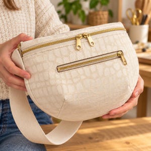 May include: A cream-colored crescent-shaped crossbody bag featuring a textured crocodile-embossed pattern. The bag includes a wide fabric strap, a main zippered compartment with two gold-tone pulls, and a smaller front zippered pocket with a gold-tone pull.