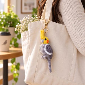 May include: A crocheted cockatiel bird keychain featuring a grey body, white wing accents, a yellow head crest, and orange cheek patches. The bird is attached to a gold-toned metal lobster clasp and ring, hanging from the strap of a cream canvas tote bag.
