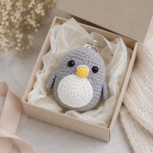 May include: A small, round, crocheted penguin keychain featuring a grey body, a white belly, a yellow beak, and two black bead eyes. The plush penguin is nestled inside a square cardboard gift box lined with white tissue paper.