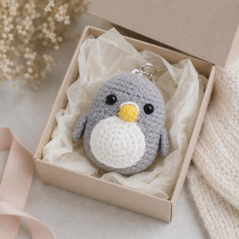 May include: A small, round, crocheted penguin keychain featuring a grey body, a white belly, a yellow beak, and two black bead eyes. The plush penguin is nestled inside a square cardboard gift box lined with white tissue paper.