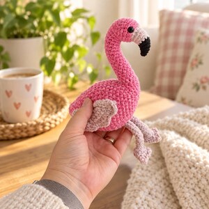 May include: A small, handcrafted crochet flamingo toy featuring a bright pink body, light pink wings, and a black-tipped beak. The amigurumi-style bird has a curved neck and dangling legs, showcasing detailed yarn stitching throughout its soft texture.