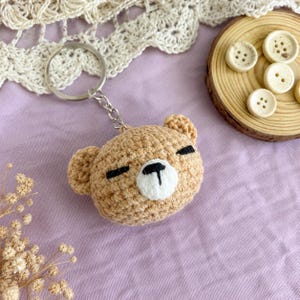 May include: A tan crochet bear head keyring featuring a white muzzle, black embroidered eyes, and a black nose. The bear is attached to a silver metal split ring and chain. The design is round with two small ears on the top sides of the head.