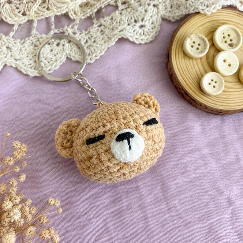May include: A tan crochet bear head keyring featuring a white muzzle, black embroidered eyes, and a black nose. The bear is attached to a silver metal split ring and chain. The design is round with two small ears on the top sides of the head.