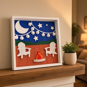 May include: A square white shadow box frame containing a layered 3D art piece. The scene depicts two white Adirondack chairs on a terracotta-colored patio with a small fire pit, set against a dark blue night sky with a white crescent moon and stars.