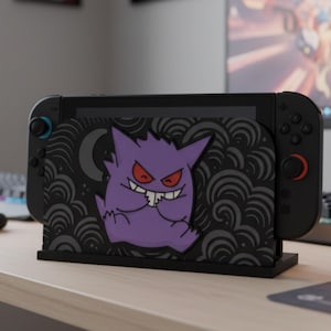 Könnte beinhalten: Schwarze Nintendo Switch-Dockingstation mit einem lila Gengar-Design mit roten Augen. Die Dockingstation hat einen schwarzen Sockel und ein dekoratives Muster aus Wirbeln und einem Halbmond. Die Switch-Konsole wird in die Dockingstation eingesetzt.