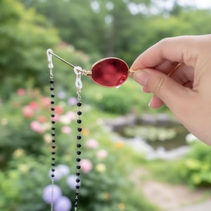 May include: Red oval sunglasses with a gold frame are held up, showcasing a black beaded eyeglass chain. The background is a blurred view of a garden with green foliage and colorful flowers.