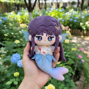 Puede incluir: Muñeca de sirena de peluche con cabello morado y cola azul. La muñeca tiene una cara detallada con ojos grandes y una expresión triste. La parte superior de la sirena es blanca con detalles rojos y aletas azules.