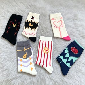 May include: Six pairs of colorful socks with various designs. One pair is black with a chicken and red crosses. Another is white with a black monster design. A third pair is white with a pink heart. The fourth pair is white with a gray top and a yellow design. The fifth pair is white with red stripes and a duck. The sixth pair is navy blue with a geometric design.