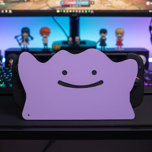 Könnte beinhalten: Eine hellviolette Ditto-förmige Nintendo Switch-Hülle mit einem lächelnden Gesicht. Die Hülle liegt auf einer schwarzen Nintendo Switch-Konsole. Der Hintergrund umfasst einen Computermonitor und Figuren.