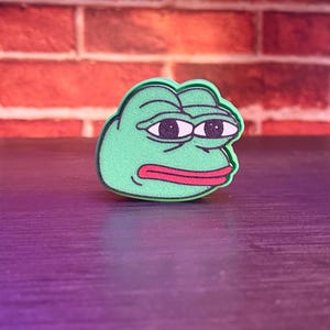 May include: A green cartoon frog face with black eyes and a red mouth, displaying a sad expression. The frog is set against a purple surface, with a red brick wall in the background.