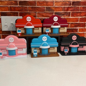 May include: Five miniature Pokémon Center building models in red, maroon, pink, blue, and black colorways. Each structure features a central dome with a Poké Ball emblem, a blue door, and a small matching console accessory placed in front.