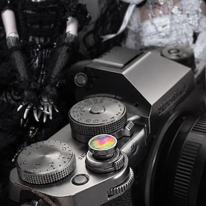 Puede incluir: Una cámara sin espejo plateada con lente negra, que presenta un botón de disparo iridiscente multicolor, un dial de compensación de exposición plateado y un dial de velocidad de obturación plateado. Se ve un enganche de correa de cuero rojo en el lateral.