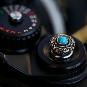 Puede incluir: Primer plano de un componente de motocicleta con una tapa decorativa plateada con un diseño ornamentado y una piedra turquesa central. La tapa está sobre un fondo negro, con un indicador visible en el fondo.