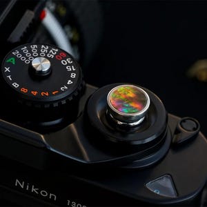 May include: Close-up of a black Nikon 1300 camera with a colorful, iridescent button. The camera's dials and settings are visible, including the ISO and shutter speed controls. The button has a silver rim and a multi-colored surface.