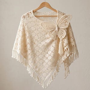 May include: Cream-colored crochet poncho with a large butterfly accent. The poncho features a geometric pattern and is trimmed with fringe and pearl-like beads. The butterfly is also crocheted and has a tassel. The poncho is hanging on a wooden hanger.