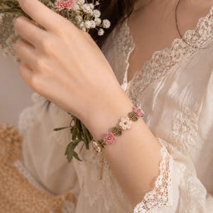 Può includere: Un braccialetto delicato caratterizzato da una serie di piccoli ciondoli a forma di fiore all'uncinetto nei toni del rosa, crema e verde. Gli elementi floreali sono collegati da componenti in metallo dorato, inclusa una catenina e una chiusura.
