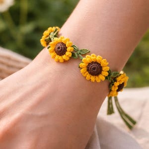 May include: Crocheted sunflower bracelet with yellow petals and brown centres. The bracelet is connected by green crocheted leaves and a green tie closure. The bracelet is worn on a wrist.