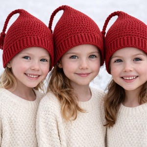 May include: Three young people wearing matching red knit hats with a long braided tassel and cream-colored sweaters. The hats have a pointed top. The people are smiling, with fair skin and blonde hair.