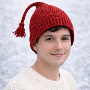 May include: A red knitted hat with a long, braided tassel and a tassel at the end. The hat is worn by a person wearing a cream-colored sweater. The hat is a winter accessory.