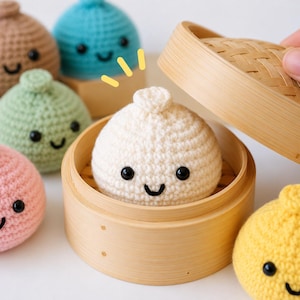 May include: A collection of six colorful crocheted dumpling toys with black embroidered eyes and smiles. One white dumpling sits inside a round bamboo steamer basket, while the others are arranged around it in pink, green, brown, blue, and yellow.