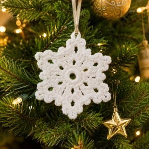 Könnte beinhalten: Ein weißes, gehäkeltes Schneeflocken-Ornament mit einer kreisförmigen Mitte und sechs spitzen Armen mit komplexen Maschen. Das Ornament ist an einer dünnen, hellen Bandschlaufe aufgehängt und zeigt ein detailliertes, strukturiertes Handarbeitsdesign.