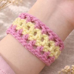 May include: A handmade crochet bracelet in pink and yellow. The bracelet is a wide band with an open, lacy design. The pink yarn forms the borders, while the yellow yarn creates the openwork pattern. The bracelet is worn on a wrist.
